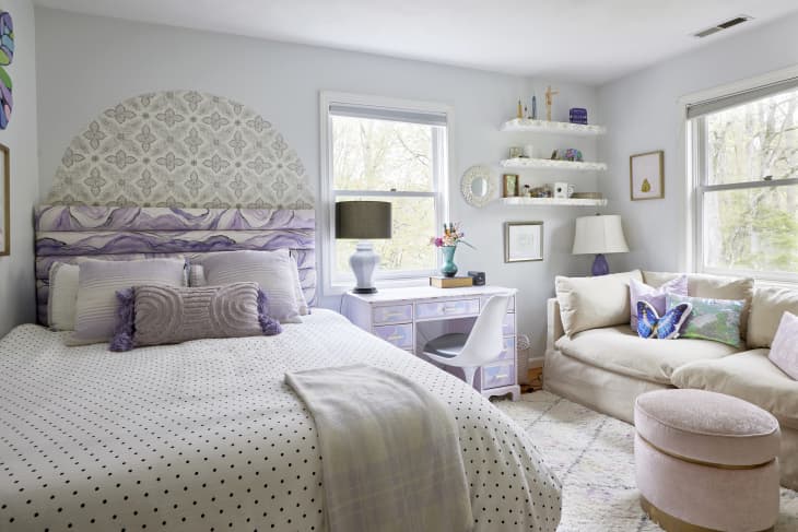 Light colored bedroom with small purple desk and polka dot covered bed. Comfortable sofa sits off to the side.