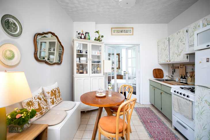 A kitchen with a round wooden table, two chairs, a white cabinet, and floral-patterned cabinets.