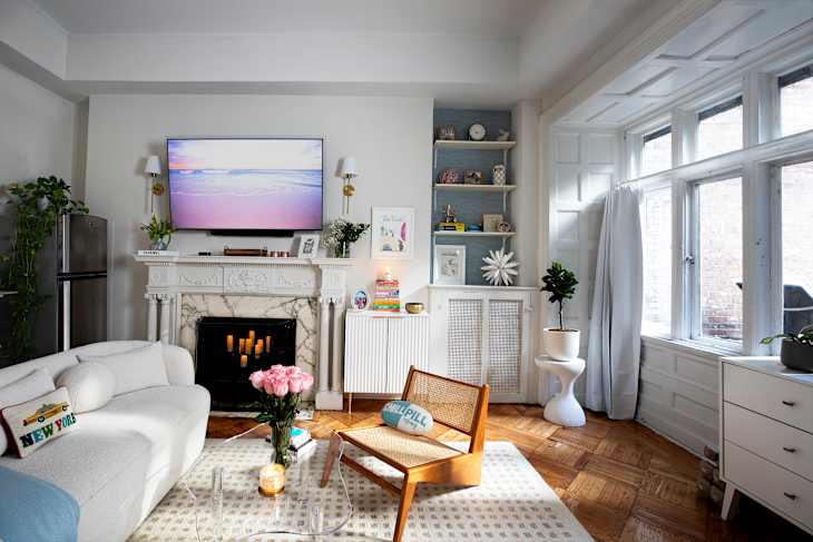 Bright living room featuring a white sofa, wooden chair, floral decor, and a marble fireplace with a TV above.