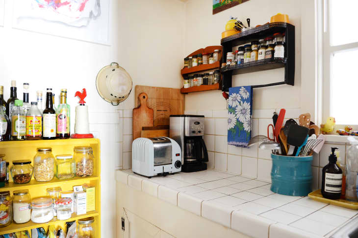 Kitchen corner with yellow shelf of bottles and jars, white toaster, coffee maker, and utensil holder on tiled counter.