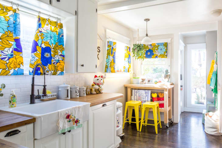 Bright kitchen with floral curtains, farmhouse sink, yellow stools, and a wooden island.