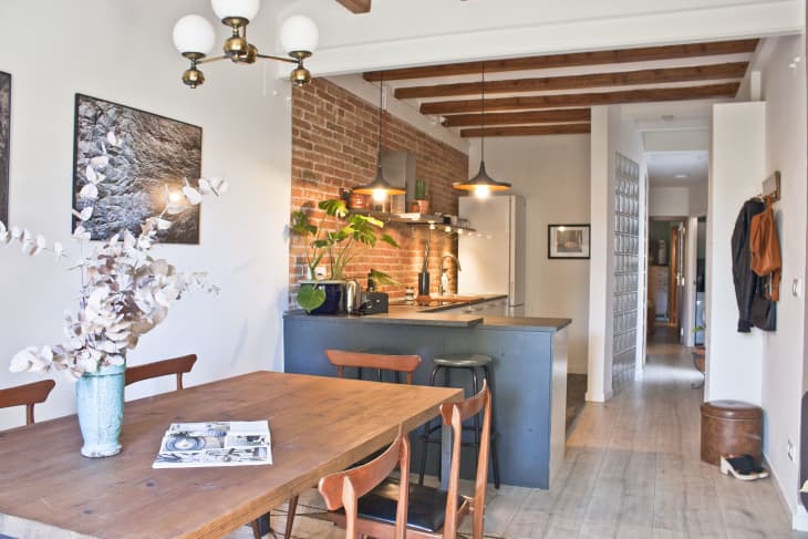 Open-plan kitchen and dining area with wooden table, chairs, brick wall, pendant lights, and plants.