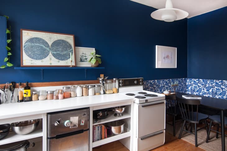 Cozy kitchen with blue walls, white shelves, vintage stove, and a small dining area with floral wallpaper and black chairs.