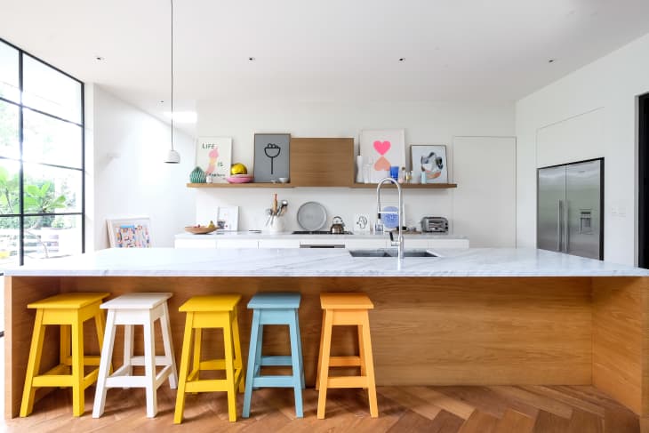 Modern kitchen with marble island, colorful stools, open shelves, and stainless steel appliances.