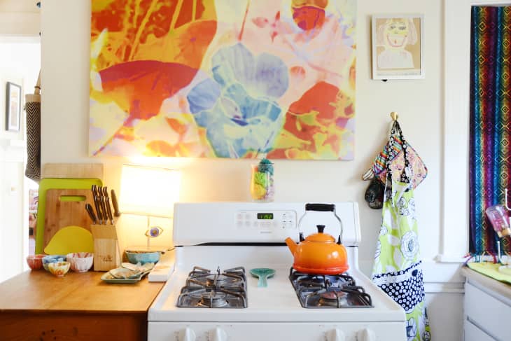 Colorful kitchen with orange kettle on stove, abstract floral art, cutting boards, and patterned aprons hanging nearby.