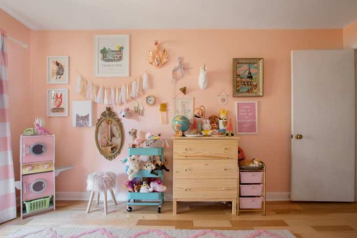 Peach-colored nursery with wall art, stuffed animals, a wooden dresser, and a pink play kitchen.