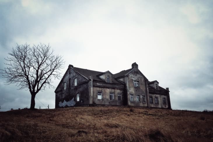 Old, weathered house on a hill with a bare tree under a cloudy sky.