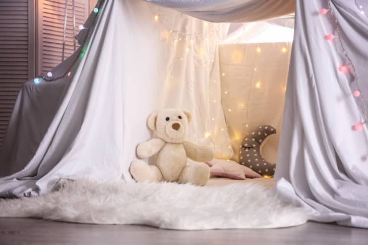 Cozy blanket fort with a teddy bear, string lights, and pillows on a fluffy rug.