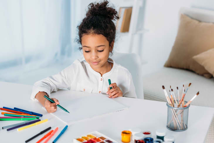 Child drawing with colored markers at a white table, surrounded by art supplies and paintbrushes in a holder.