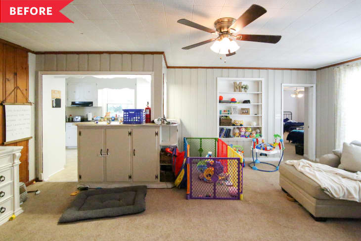 Living room with beige walls, ceiling fan, playpen, toys, and a beige sectional sofa. Kitchen visible through doorway.
