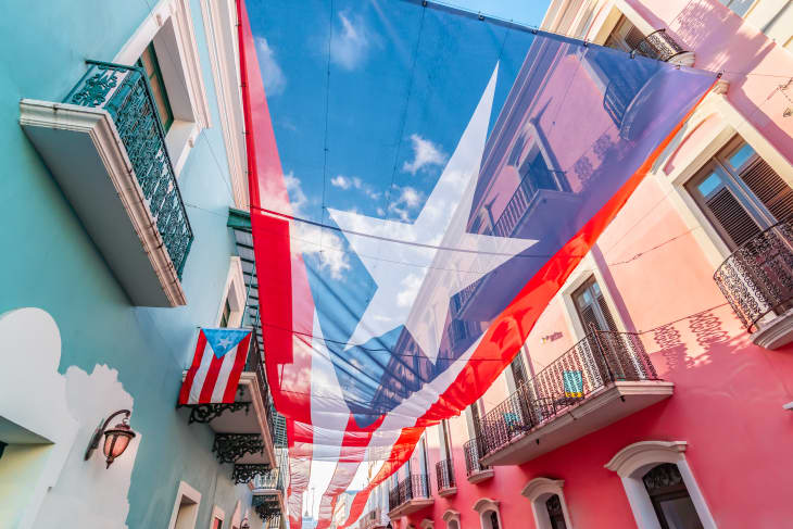 Large Puerto Rican flag hanging between colorful buildings with balconies in a narrow street.