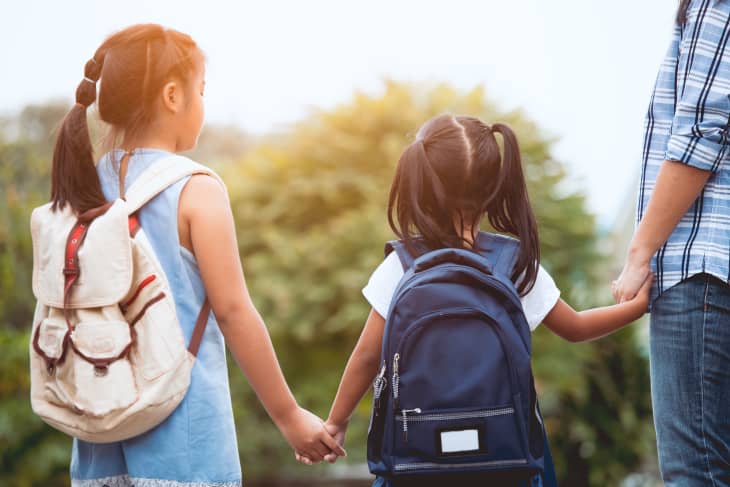 Two children with backpacks holding hands with an adult, standing outdoors.