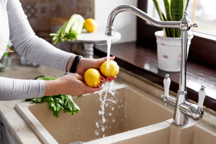 Washing two lemons under a kitchen faucet with celery and a potted plant nearby.