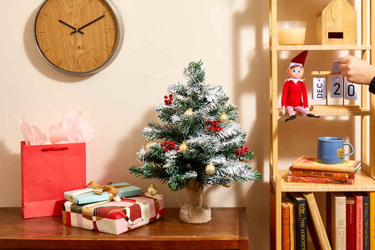 Head on shot of a living room scene with a light wood shelving unit, and dark wood sideboard to the left of it.  On the wall theres a silver and wood faced clock.  On the sideboard, theres a decorated table top tree that's been flocked, and an assortment of gifts next to it.