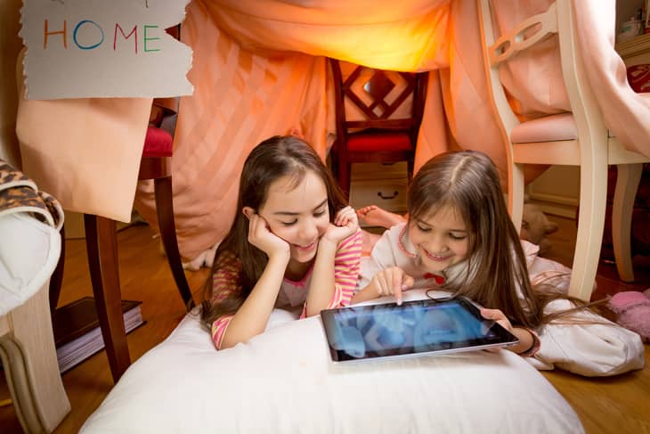 Two girls lying on pillows in a blanket fort, using a tablet.