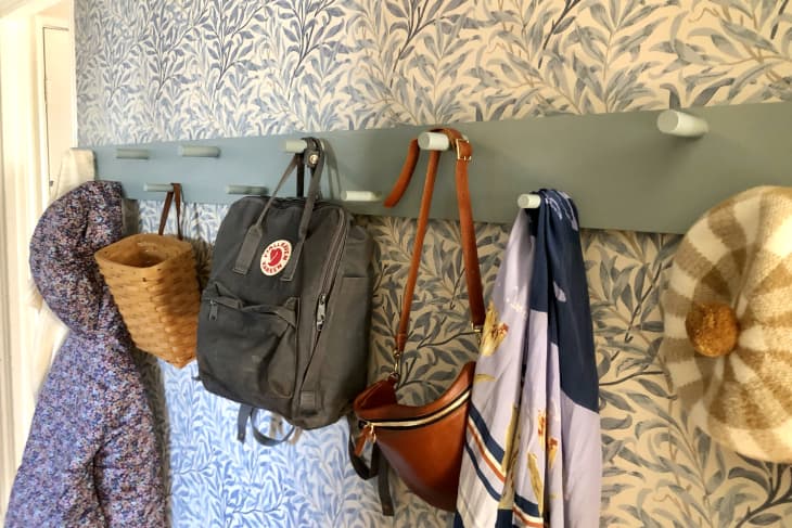 Wallpapered hallway/entryway with green wooden wall hooks. There are multiple backpacks, bags, a hat, and a floral coat hanging on them