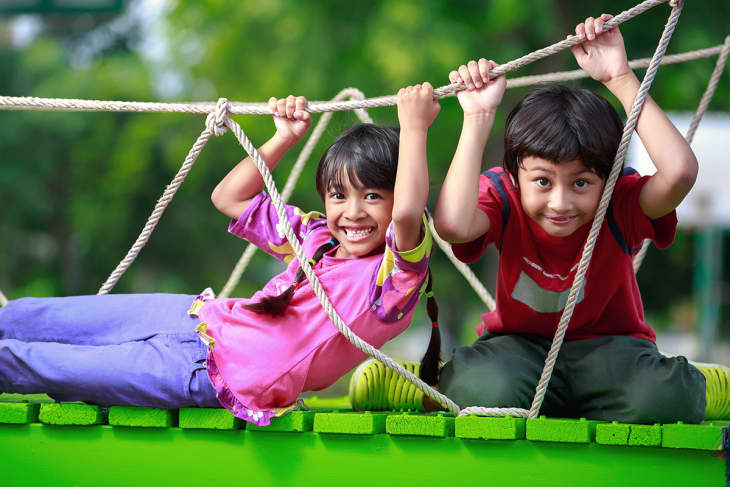 Two children smiling and playing on a rope bridge in a park, surrounded by greenery.