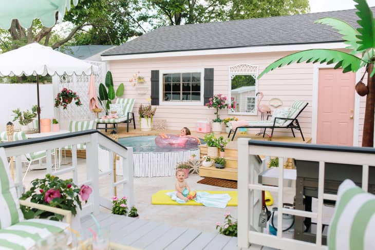 Backyard with pink pool house, lounge chairs, inflatable pool, and tropical decor. Child on yellow mat, woman in pool.