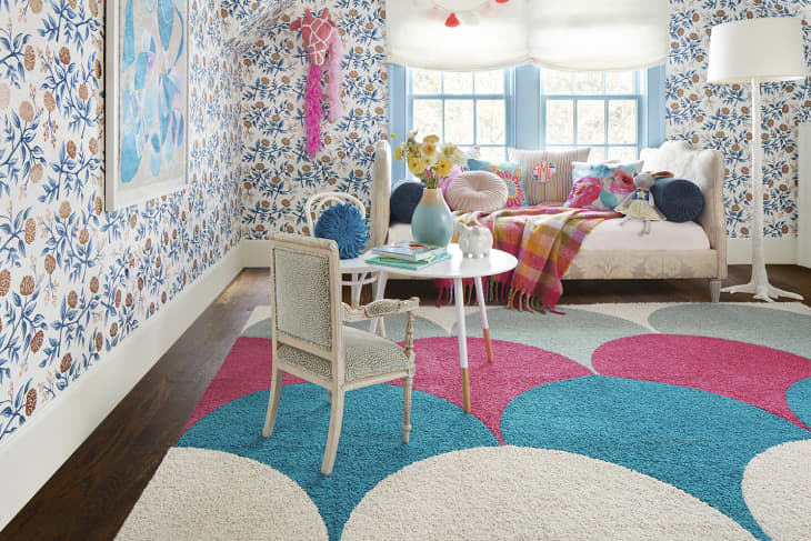 Colorful playroom with floral wallpaper, patterned rug, daybed with pillows, and a small table with books and a vase.