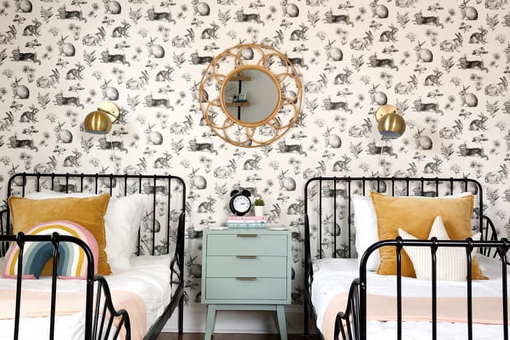 Two twin beds with mustard pillows, rabbit wallpaper, gold sconces, and a mint nightstand with a round mirror above.