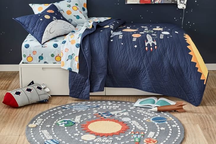 Space-themed kids' bedroom with navy bedding, solar system rug, and books on a shelf.
