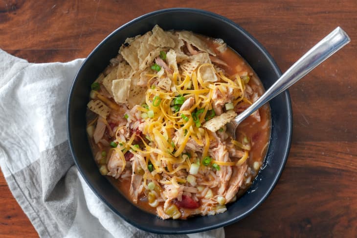 Bowl of chicken tortilla soup topped with shredded cheese, tortilla chips, and green onions on a wooden table.