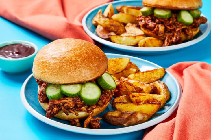 Pulled pork sandwiches with cucumber slices and potato wedges on blue plates, accompanied by a small bowl of sauce.