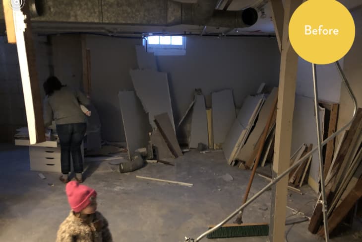 Unfinished basement with drywall sheets, exposed pipes, a woman organizing, and a child in a pink hat.