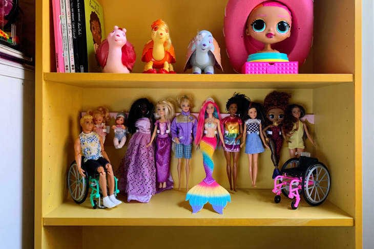 Diverse dolls on a yellow shelf, including a mermaid, dolls in wheelchairs, and various outfits.