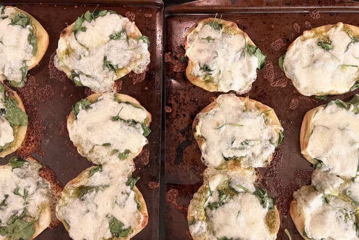 Mini pizzas topped with melted cheese and spinach on two baking sheets.