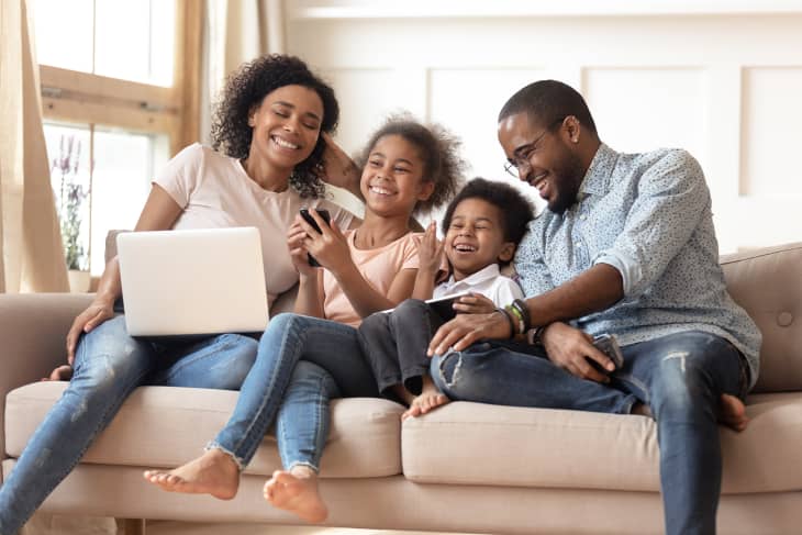 Black family of four sitting on couch smiling and interacting. Mother, adolescent daughter, preschool-aged boy, and father are interacting with screens while also talking to one another.