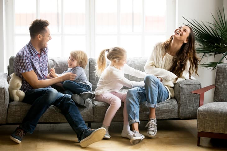 Family of four playing on the couch in front of a bright window. Dad and son turn towards each other, while daughter leans over mother, who is smiling.