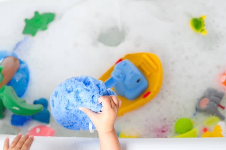 Child playing with blue sponge in a bubble bath with yellow toy boat and colorful bath toys.