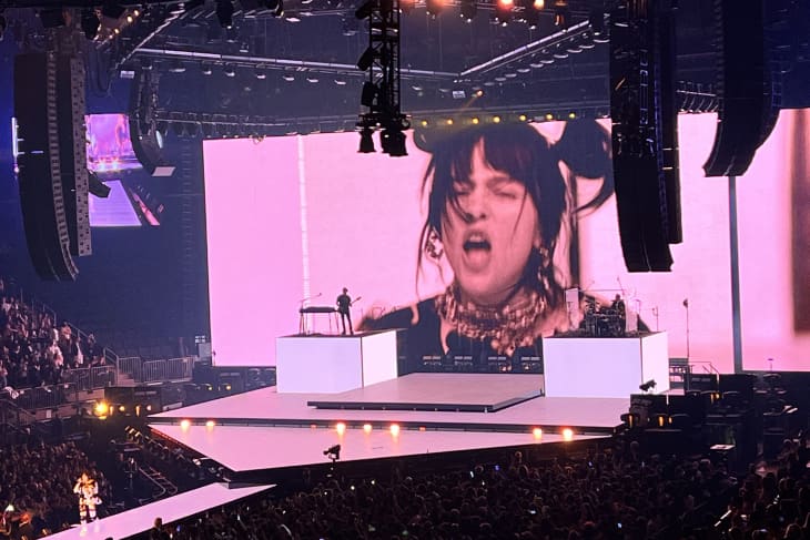 Concert stage with large screen displaying performer, surrounded by audience and vibrant lighting.