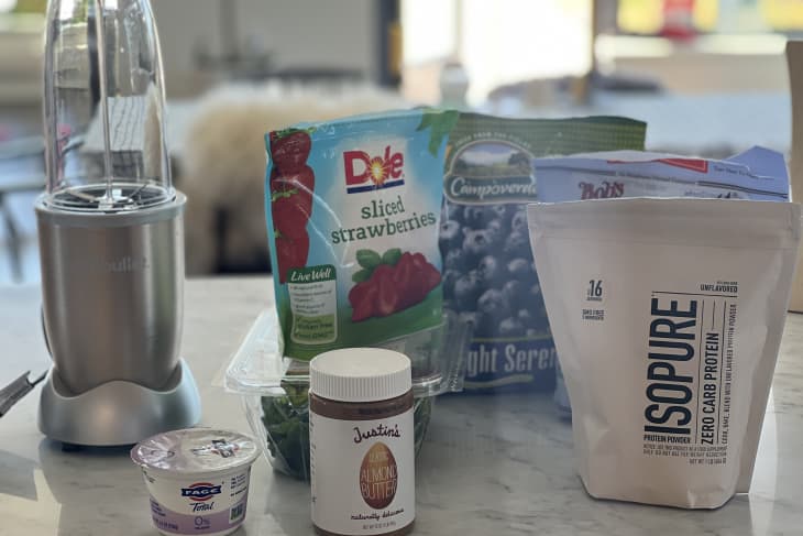 Blender with yogurt, almond butter, sliced strawberries, blueberries, and protein powder on a kitchen counter.