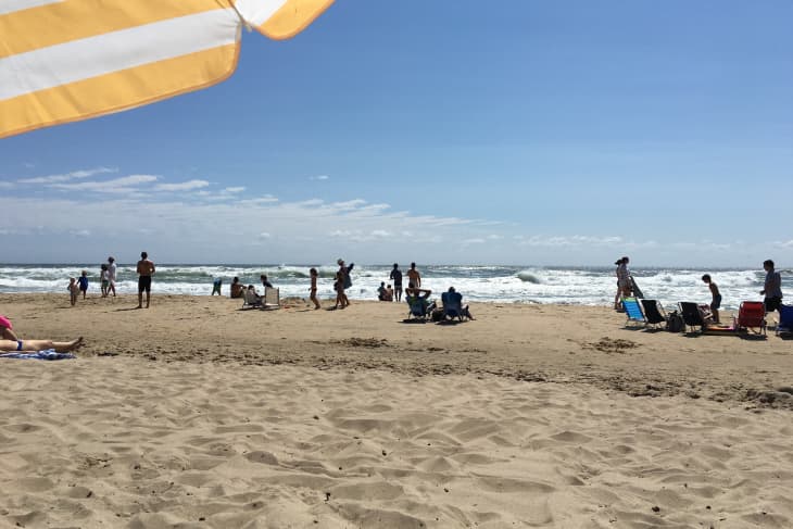 People enjoying a sunny day at the beach with waves in the background and a striped umbrella in the foreground.