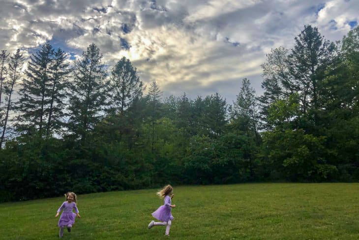 Girls in purple dresses running outside in a green field against tall evergreen trees and a cloudy sky