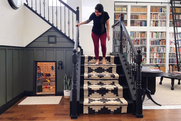 Woman descending stairs with patterned carpet, child visible in play area under stairs, bookshelves in background.