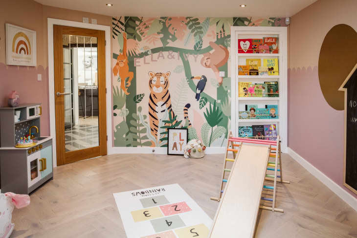 Playroom with painted mural with kids names, tiger, toucan, sloth, monkey. Bookshelf with books arranged by color. kids play kitchen and wood slide