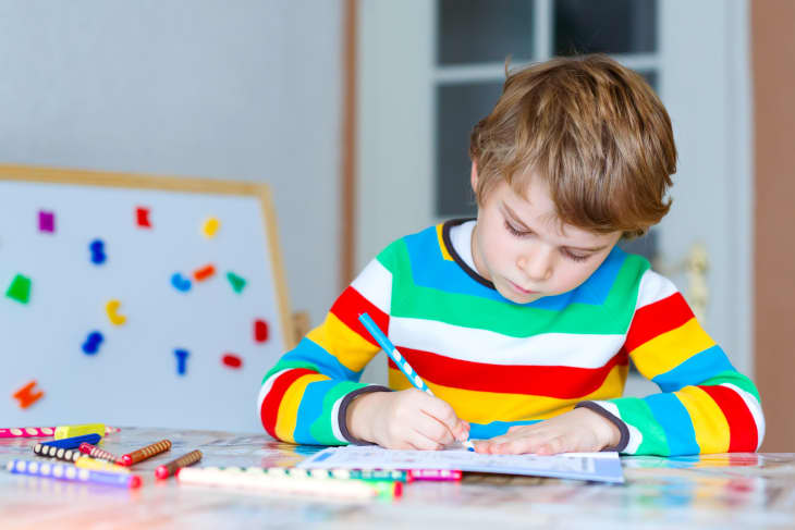 Child in a rainbow-striped shirt drawing with colored pencils at a table, magnetic letters on a board in the background.