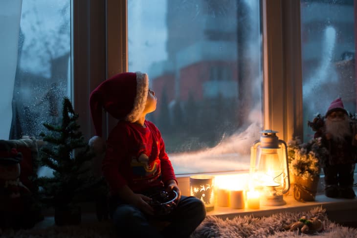 Kid in santa hat next to a lantern looking outside the window at dusk