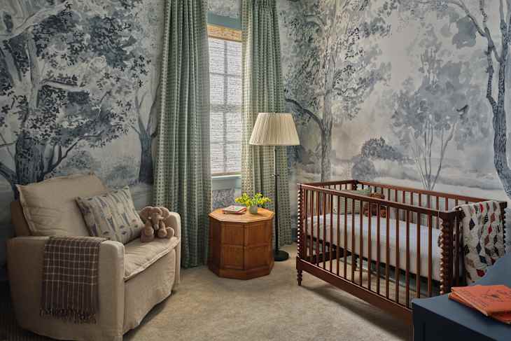 Nursery with a wooden crib, beige armchair, green curtains, and nature-themed wallpaper featuring trees.