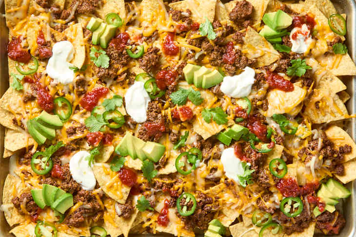overhead shot of nachos in a sheet pan,  topped with cilantro, avocado, sour cream and salsa.