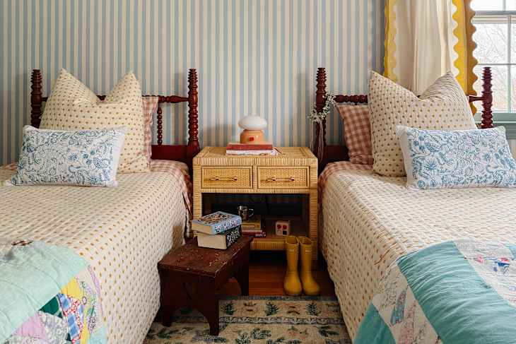 Two twin beds with patterned quilts, decorative pillows, a rattan nightstand, and yellow rain boots on a rug.