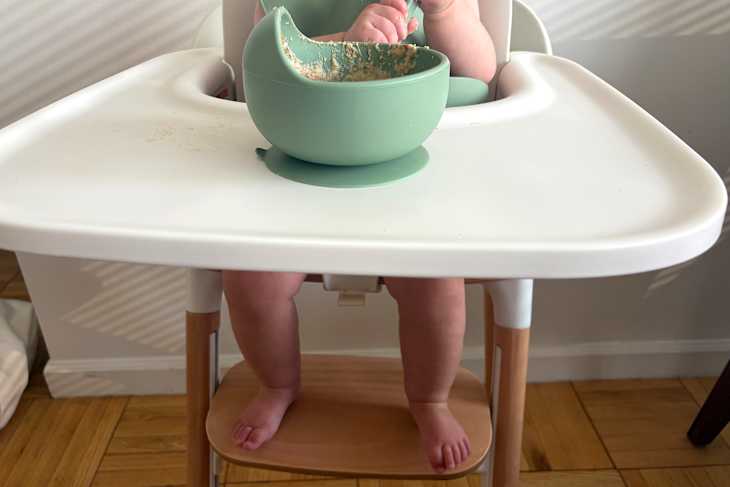 A baby in a high chair playing with a green bowl of food, with bare feet visible on the wooden footrest.
