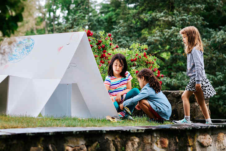 Three children playing near a white cardboard playhouse in a garden with red flowers.