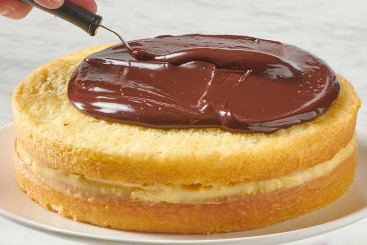 a whole boston cream pie on a cake stand being topped with chocolate ganache