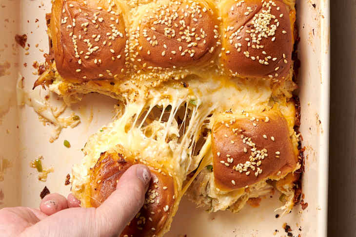 overhead shot of chicken sliders in a baking sheet; a hand pulls one away from chicken slider group showing cheese pull