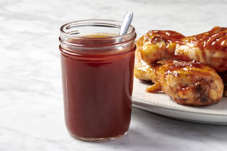 head on shot of a jar of honey barbecue sauce with a silver spoon on it and a white plate of chicken behind it to the right hand side both placed on a marble surface