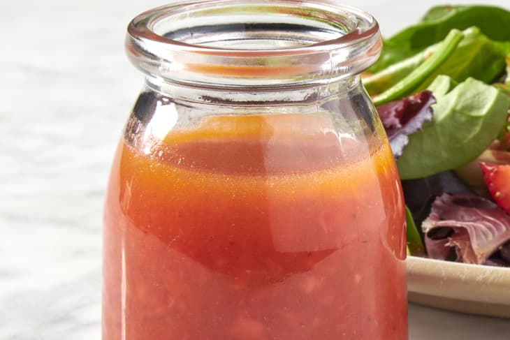 a head on shot of strawberry vinaigrette in a jar placed on a marble surface with a plate of salad behind the jar
